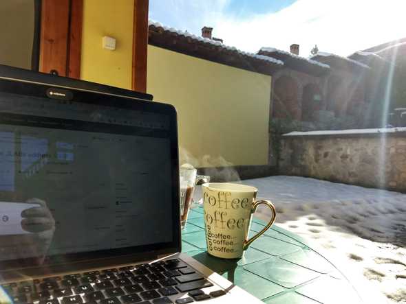 Laptop and coffee mug on a table in the terrace. Lawn covered of snow in the background.